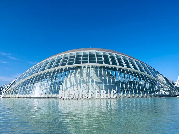 Alt-text: The Hemisfèric building with a curved glass structure reflecting in water, located in the City of Arts and Sciences in Valencia, under a clear blue sky.