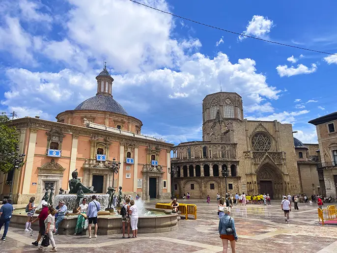 Alt-text: The Plaza de la Virgen in Valencia, featuring the ornate Neptune Fountain, historic buildings, and groups of people under a partly cloudy sky.