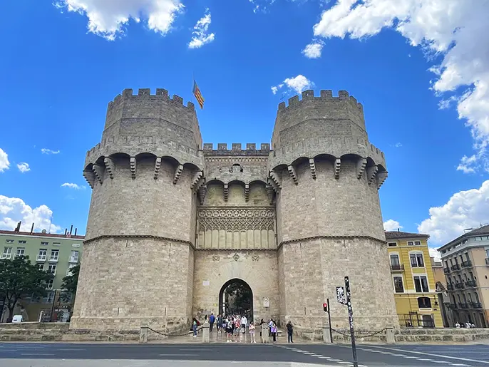 Alt-text: The historic Torres de Serranos in Valencia, a large stone gateway with two tall towers, under a partly cloudy blue sky with people gathered at the entrance.