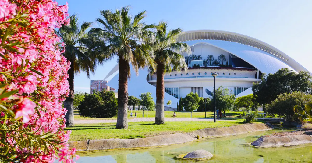 A scenic view of Turia Park in Valencia featuring palm trees, blooming pink flowers, a modern architectural building, and a cyclist in the background, showcasing a peaceful spot for a Valencia Bike Tour.